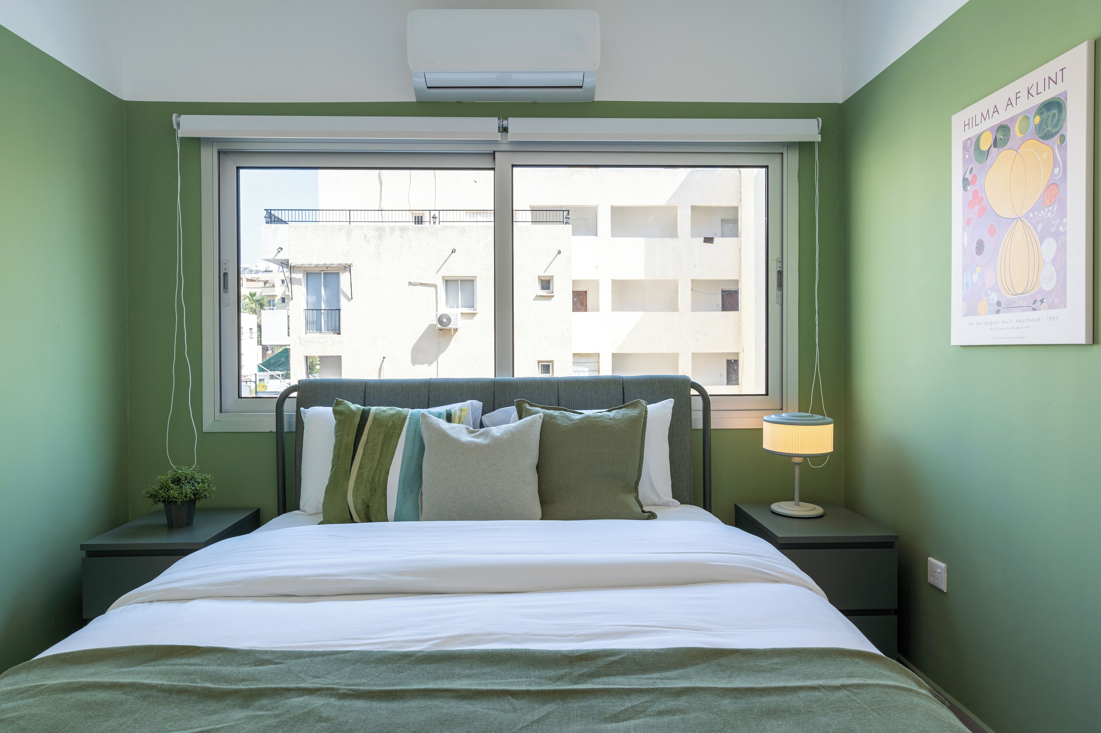 Een slaapkamer met een bed, nachtkastje, lamp, raam en een wandairconditioner.