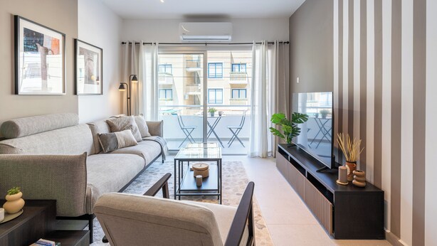 A modern living room with a sofa, a coffee table, and a TV. There is a balcony with a view of buildings and a plant on the side. - Hazel 2-BR Apt. & Balcony in Larnaca (Larnaca)