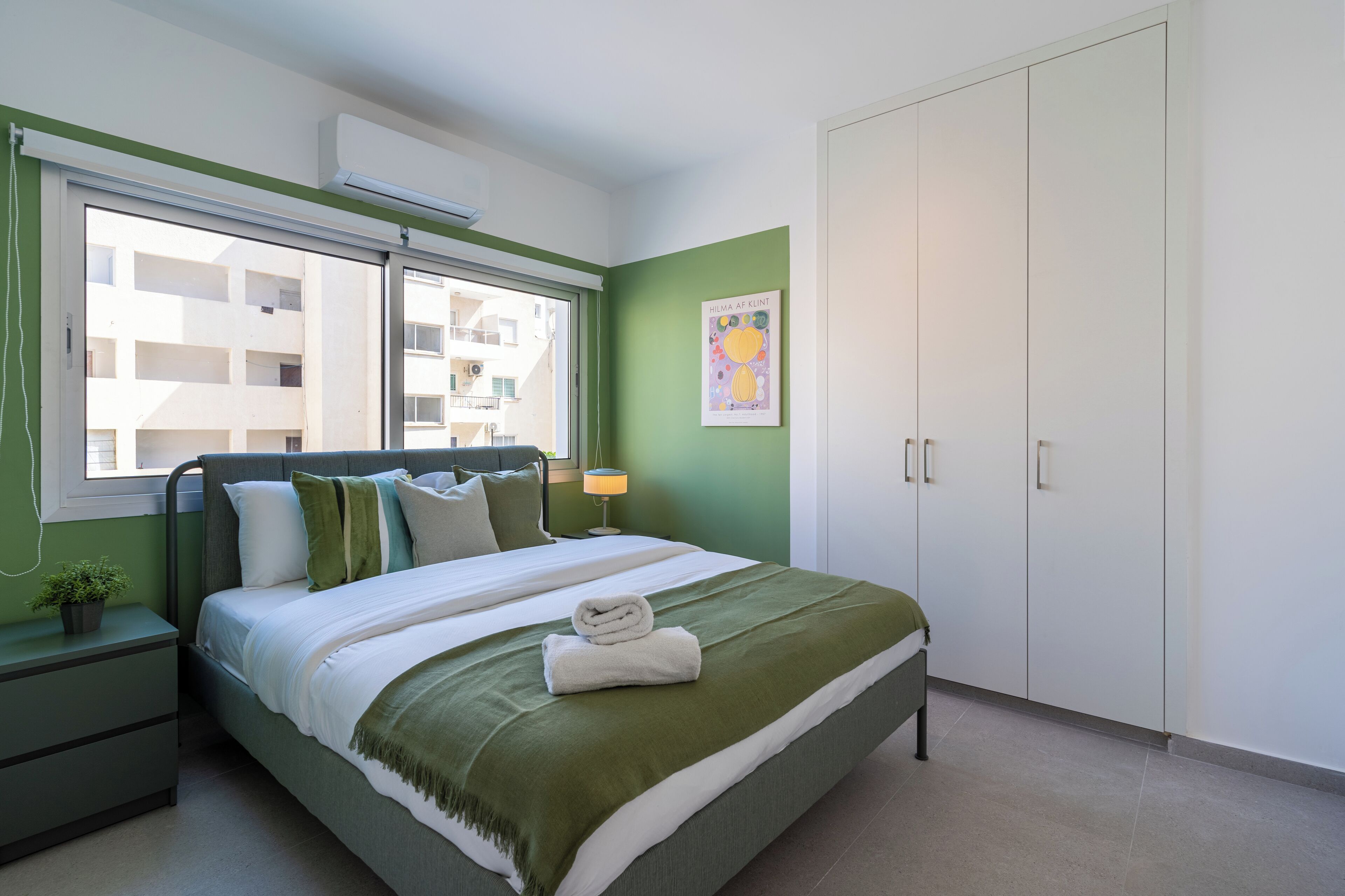 A bedroom with a bed, green walls, a window, a bedside table with a lamp, and a wardrobe.