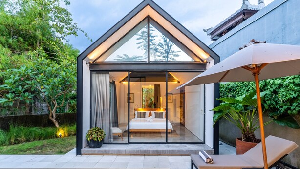 Een moderne slaapkamer met glazen wanden, een bed, een klein zithoekje en een ligbed aan het zwembad met een parasol.