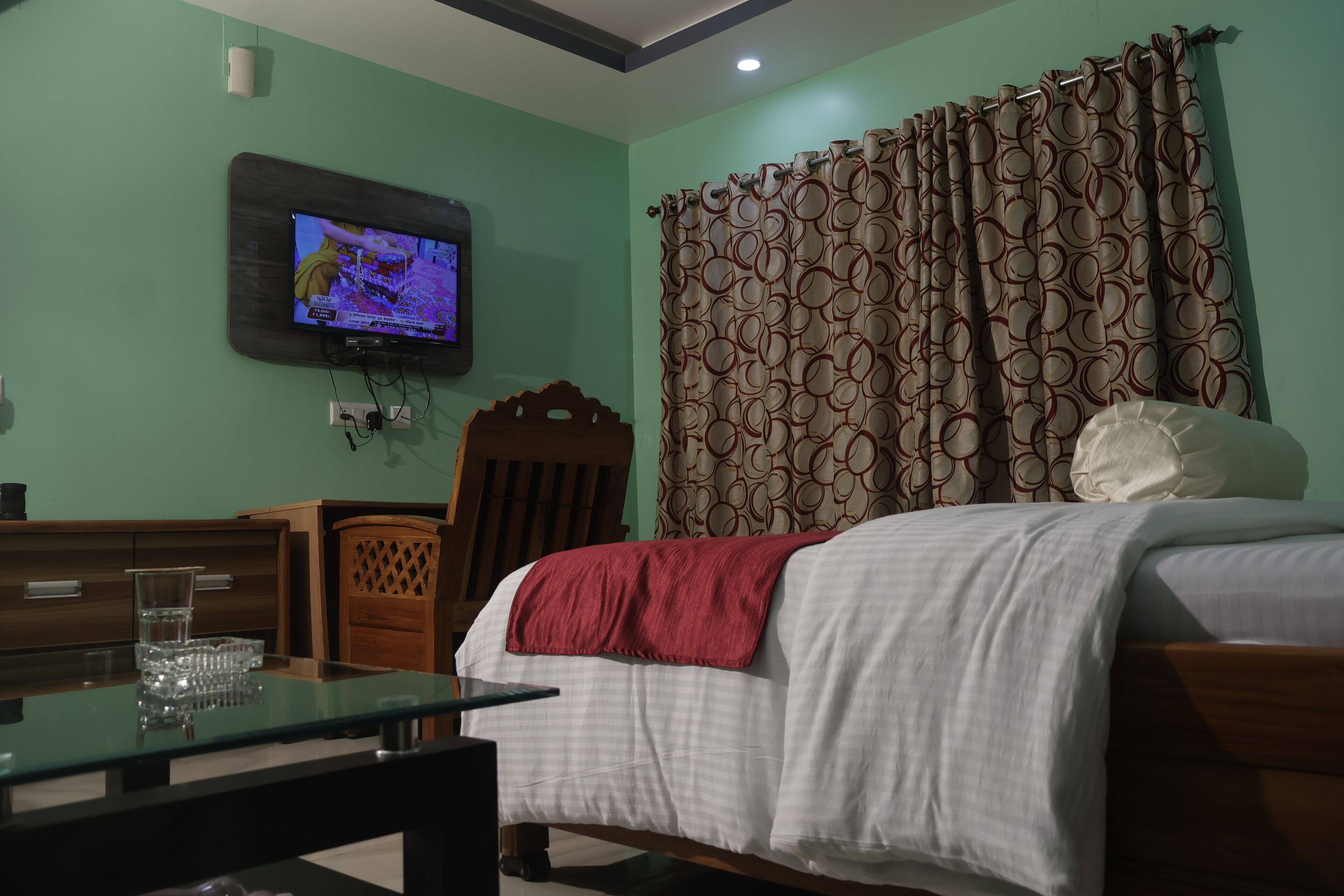 A bedroom with a bed, a television mounted on the wall, a wooden chair, a glass table with a glass set, and a patterned curtain.
