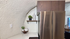 Une cuisine dotée d’un réfrigérateur en acier inoxydable, d’un plan de travail blanc, d’une étagère en bois ornée d’une plante en pot et d’un mur à texture.