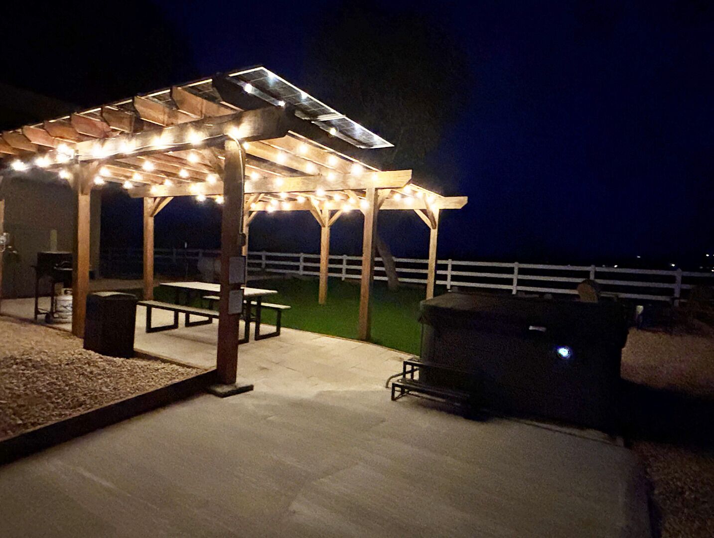 Un patio al aire libre con una pérgola de madera, luces colgantes, mesas de picnic y una parrilla.