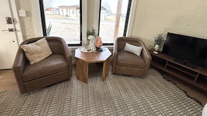 Un salon comprenant deux fauteuils en cuir, une petite table en bois ornée d’objets décoratifs, un téléviseur posé sur un meuble en bois, et de grandes fenêtres donnant sur l’extérieur.