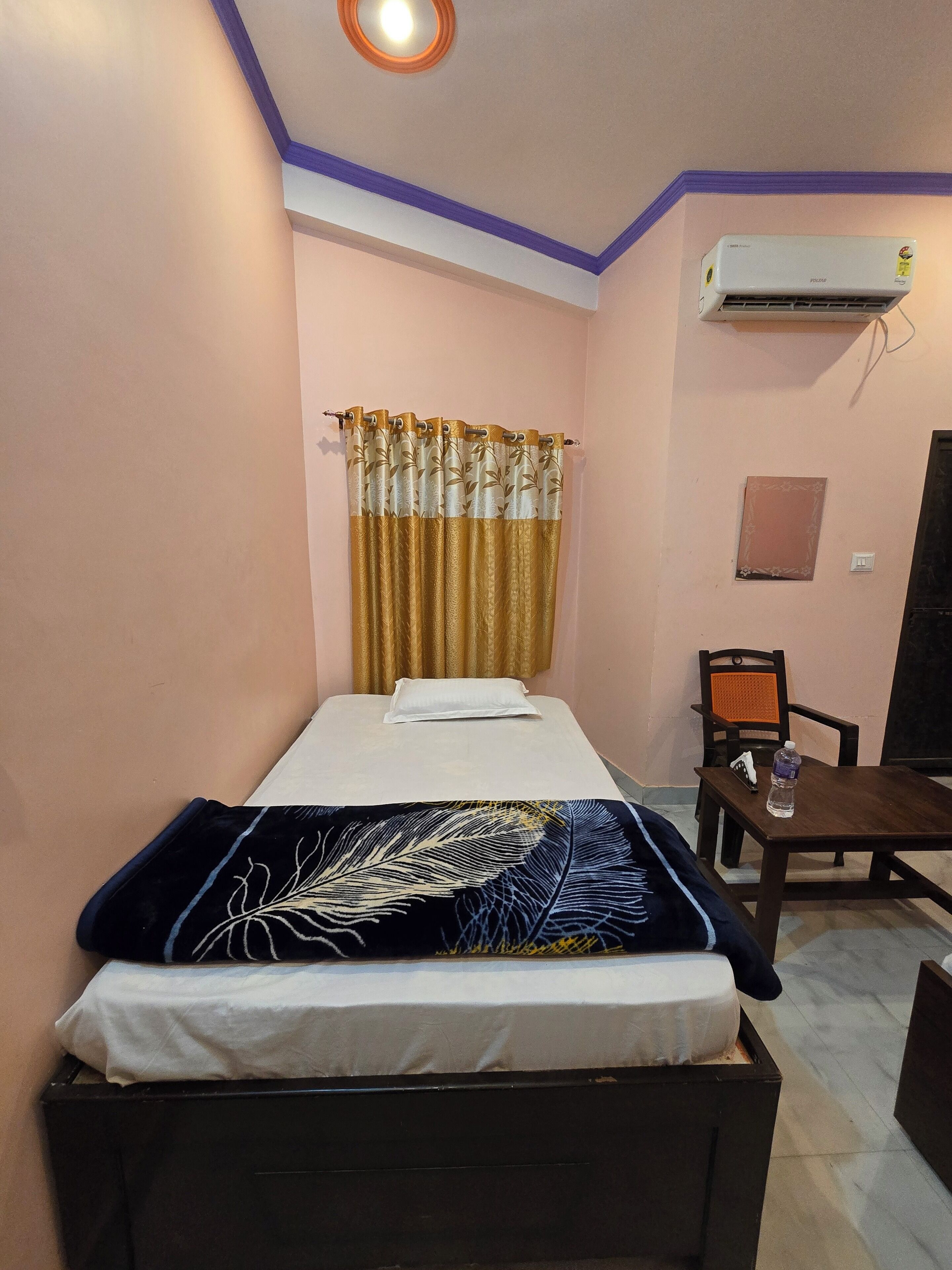 A single bed with a white mattress and a black and blue feather-patterned blanket. There is a small table with a water bottle and a chair. The room has a pink wall, a window with gold curtains, and an air conditioning unit.