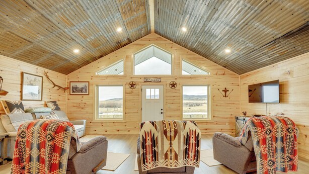 A log cabin interior with a high ceiling, two sofas with blankets, a television, and a door labeled "BUNKHOUSE". - New-Build, Mtn-View White Sulphur Springs Home! (White Sulphur Springs)