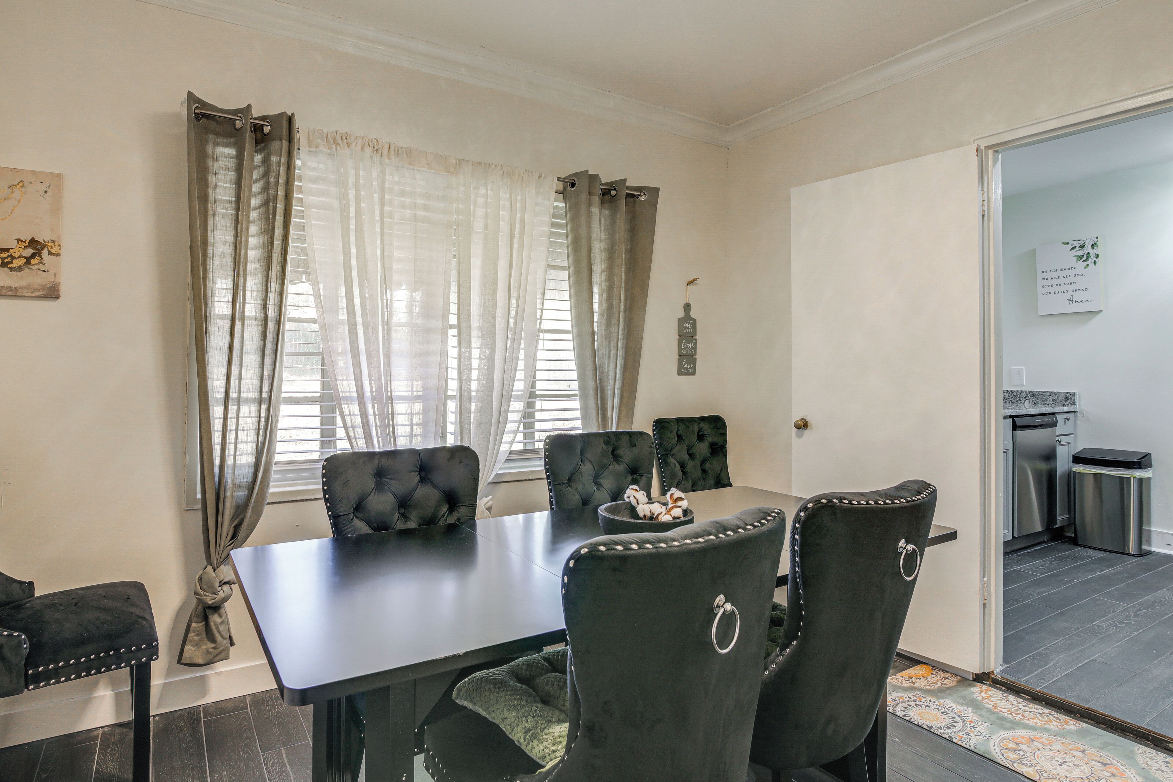 Un coin repas avec une table sombre et des chaises assorties, une fenêtre ornée de rideaux légers et une vue sur la cuisine.