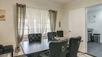 Un coin repas avec une table sombre et des chaises assorties, une fenêtre ornée de rideaux légers et une vue sur la cuisine.