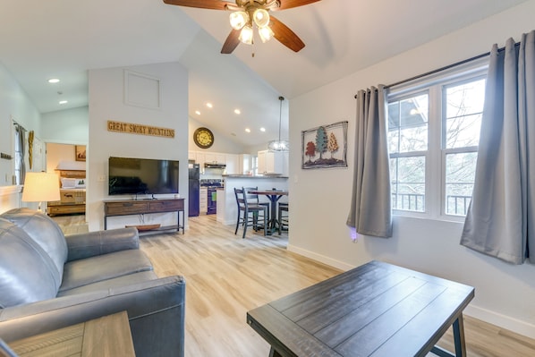 A modern living room with a leather sofa, a wooden coffee table, a TV, a ceiling fan, a kitchen in the background, and a dining area.