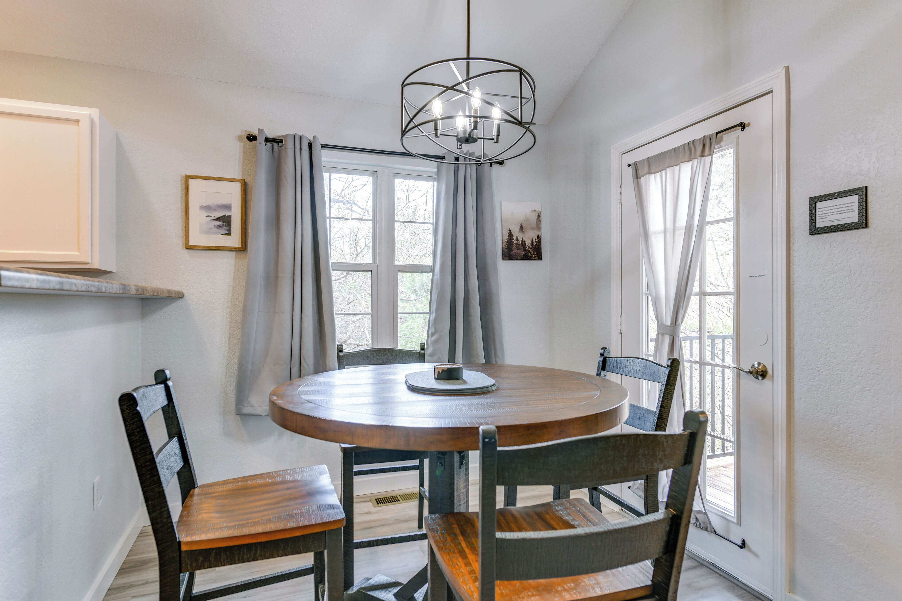 A dining area with a round table, four chairs, a chandelier, and two windows with curtains.