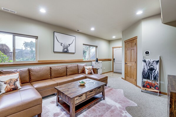 A spacious living room with a brown leather sectional sofa, a wooden coffee table, and a large window providing a view of the outside. - Near Ferry, Shops & Eats! Anacortes Townhome (Anacortes)