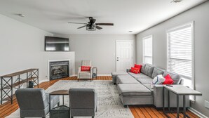 Een moderne woonkamer met een hoekbank, een open haard, een plafondventilator en een groot raam met jaloezieën.