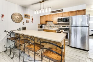 A modern kitchen with wooden cabinets, a stainless steel refrigerator, a large island with bar stools, and a wall clock.