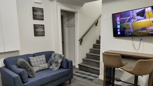 A living room with a blue sofa, two chairs, a TV displaying a show, and a staircase. - MegaVille: Serene, peaceful and relaxing apartment while working or on vacation. (Saskatoon)