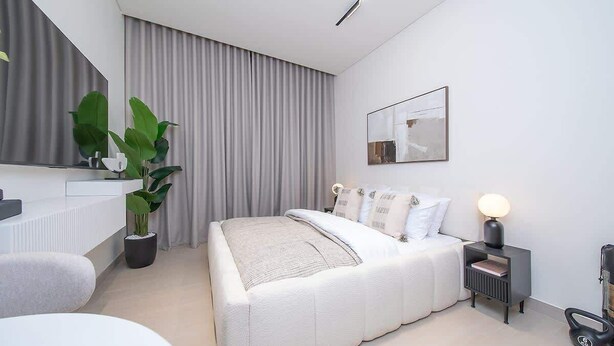 A modern bedroom with a bed, bedside table, lamp, and a potted plant. - Modern Studio in Binghatti Azure WPool & Gym (Dubai)