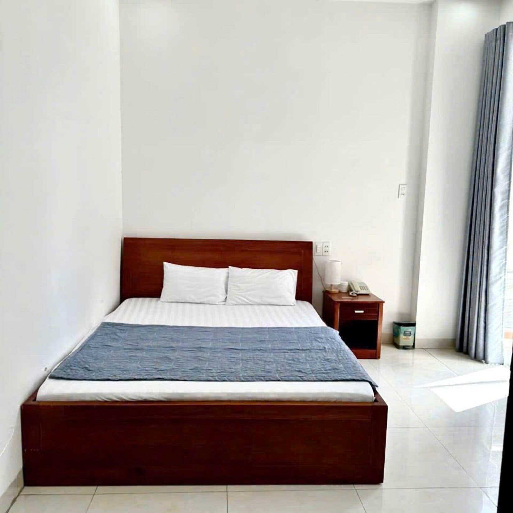 Una habitación de hotel con una cama de madera, ropa de cama blanca, una mesita de noche con lámpara y un pequeño cubo de basura.