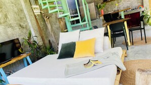 A bed with a white mattress, green and yellow pillows, and a blanket. There is a TV on a wooden stand, a plant, and a desk with a chair in the background.