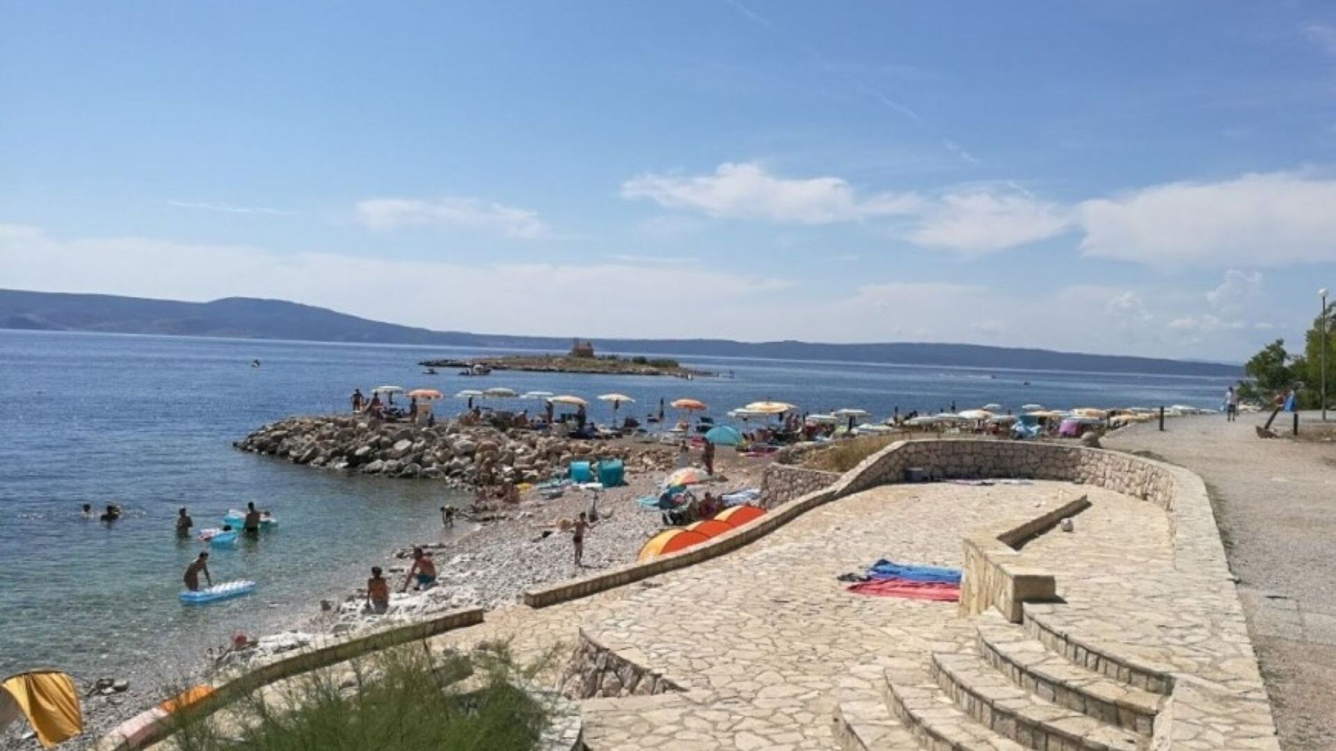 A coastal beach with a stone wall, umbrellas, and people swimming and sunbathing.