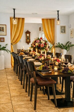 A formal dining room with a long table set for a meal, featuring a large floral centerpiece, candles, and a clock on the wall.