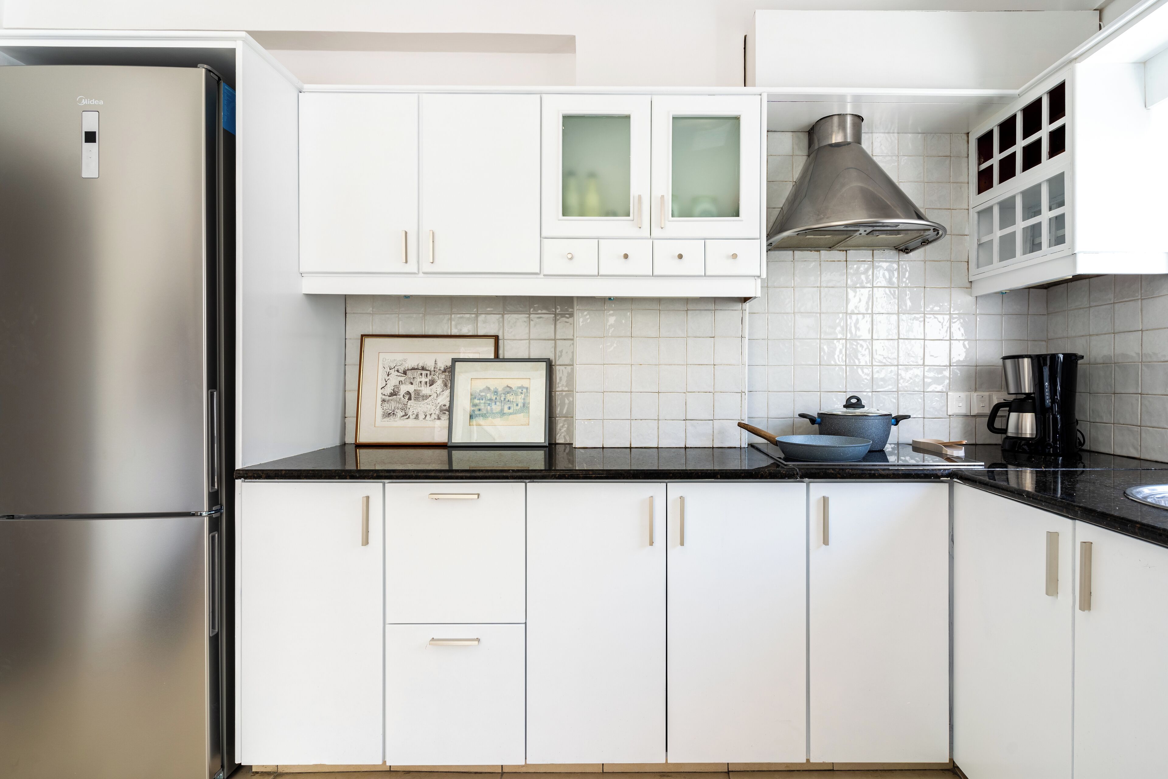 A modern kitchen with white cabinets, a stainless steel refrigerator, a black countertop, and a hanging light fixture.