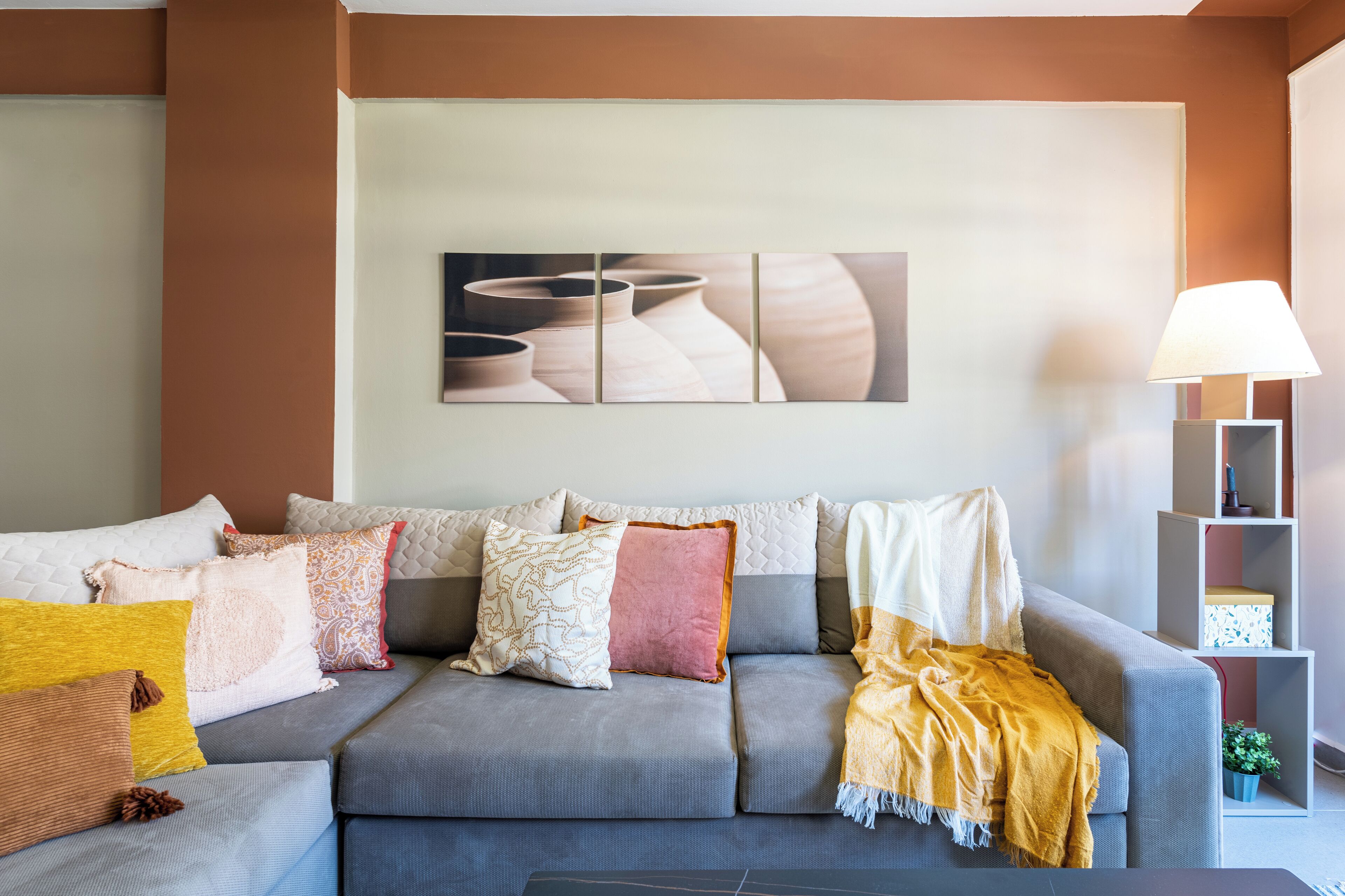 A living room with a grey sofa, multiple cushions, a wall-mounted artwork, and a side table with a lamp.
