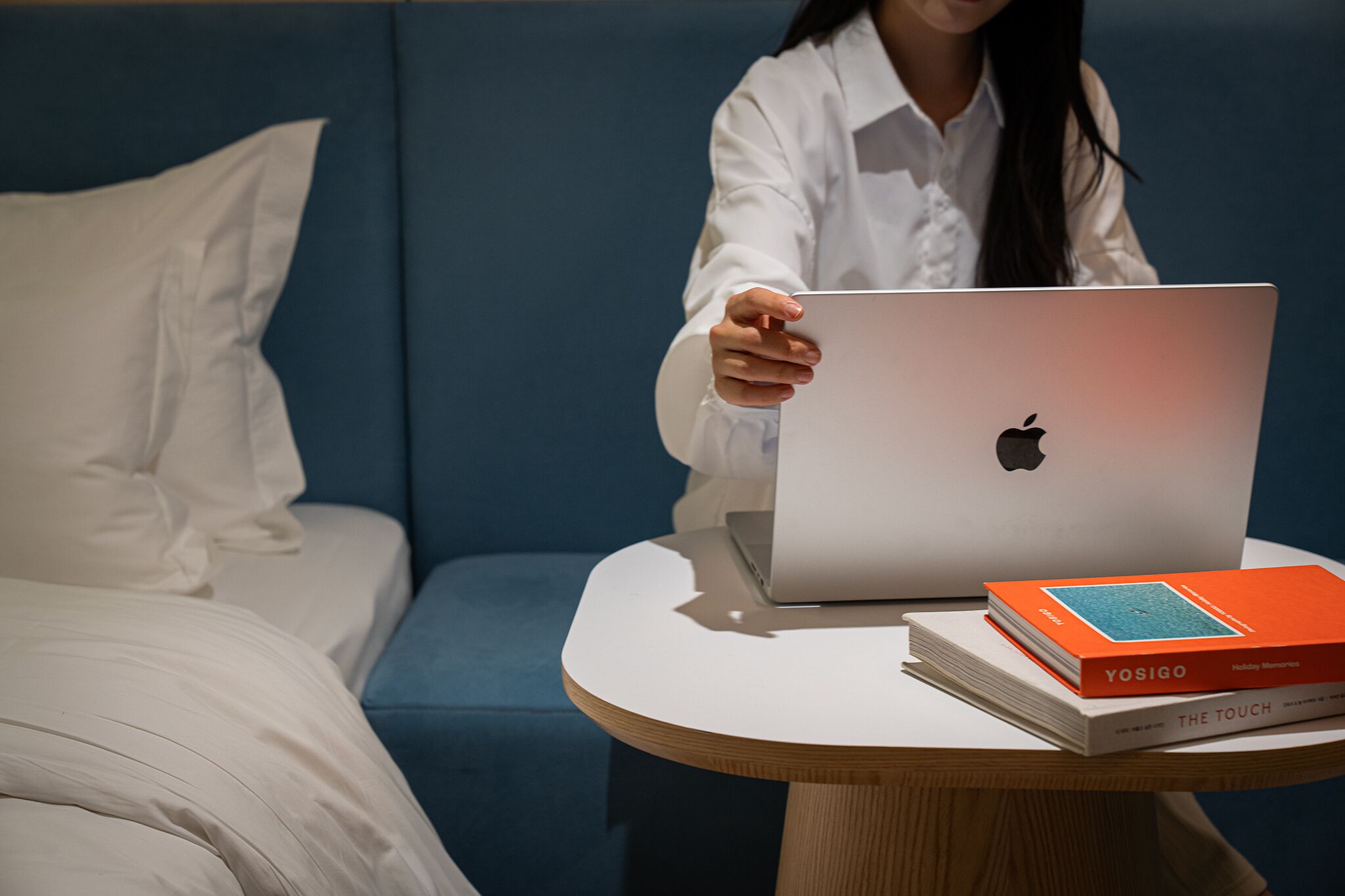 Een persoon zit op een blauwe bank met een laptop en boeken op een ronde tafel.