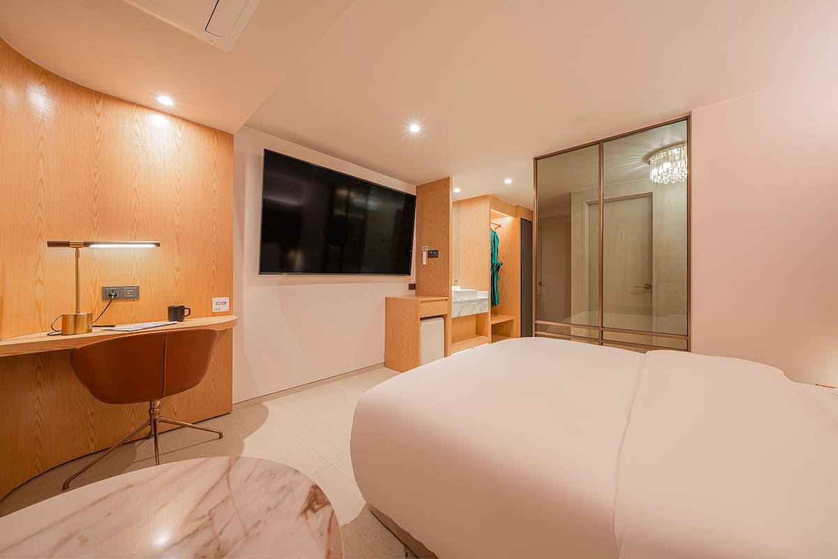 A modern hotel room with a large bed, a desk with a chair, a vanity with a mirror, and a shower area.