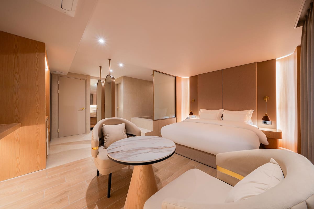 A modern hotel room with a large bed, a round table, two armchairs, and a mirror.