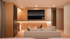 A modern hotel room with a large flat-screen TV mounted on the wall, a sofa, a small side table with a bottle and glasses, and wooden paneling. - Villa de Boni (Seoul)
