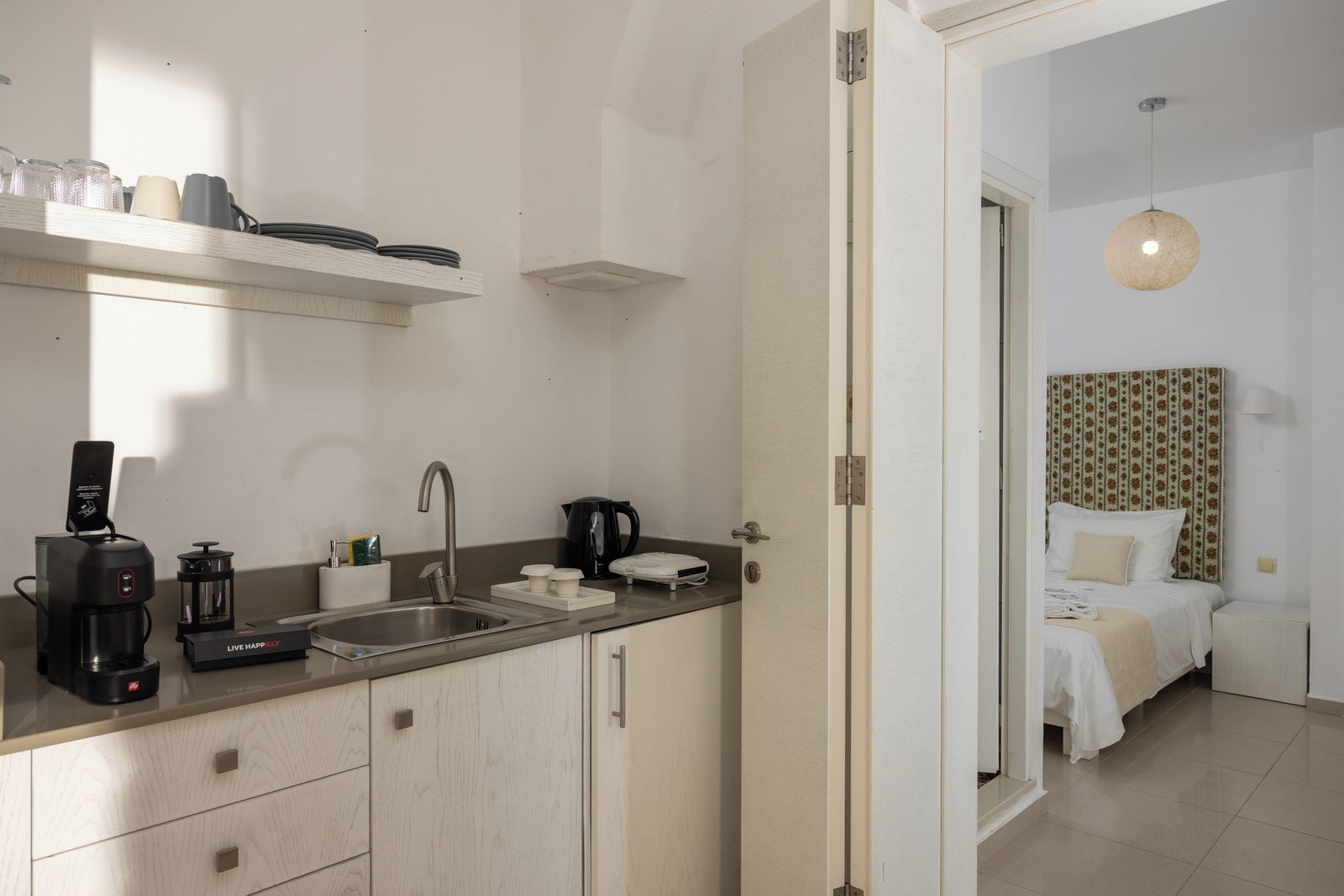 A compact hotel room with a kitchenette, a bed with patterned headboard, and a hanging light fixture.