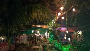 An outdoor patio area with tables and chairs, surrounded by greenery and illuminated by string lights. - River Retreat Kolad (Kolad)