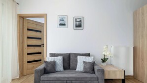 A modern living room with a grey sofa, two framed pictures on the wall, a wooden door, and a white radiator. - Elegant Apartment | Garden, Desk, Parking (Gliwice)
