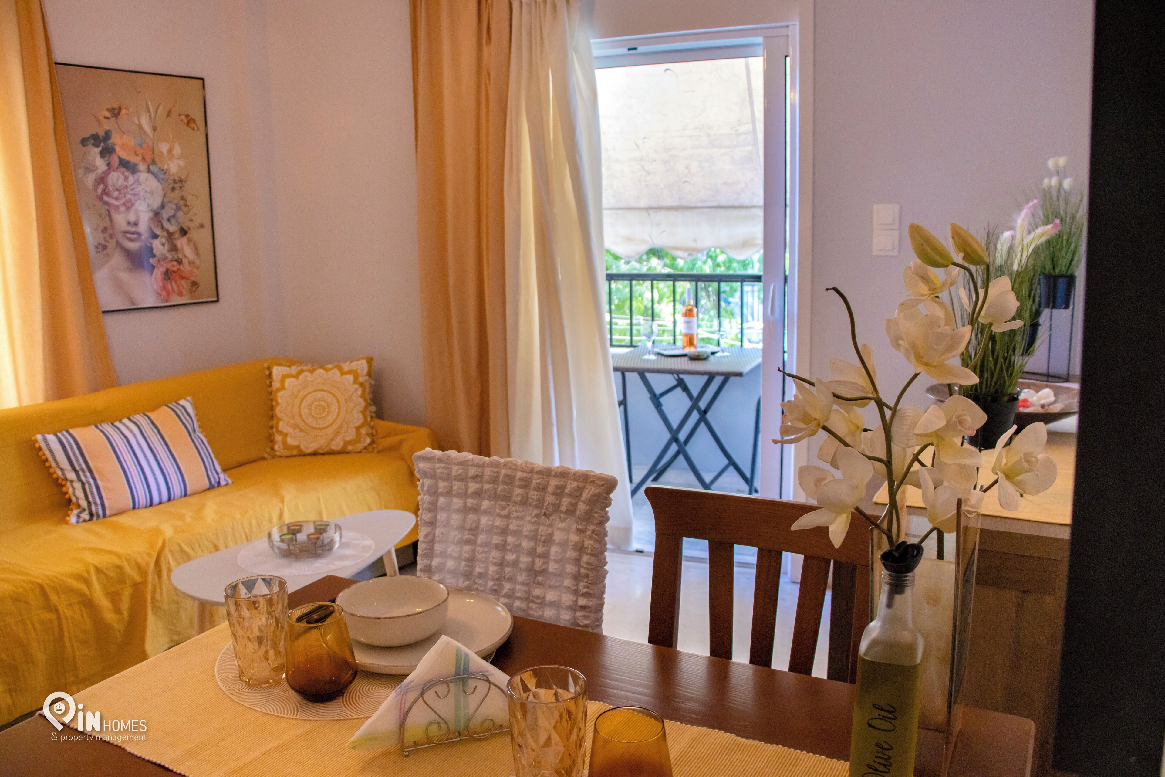 Un salon chaleureux avec un canapé jaune, une table à manger agrémentée d’un vase de fleurs et un balcon avec une table et des chaises.