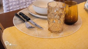 Et spisebord med en dug, en skål, et glas og en flaske. Der er en stol med en pude, et ur på væggen og et spejl.