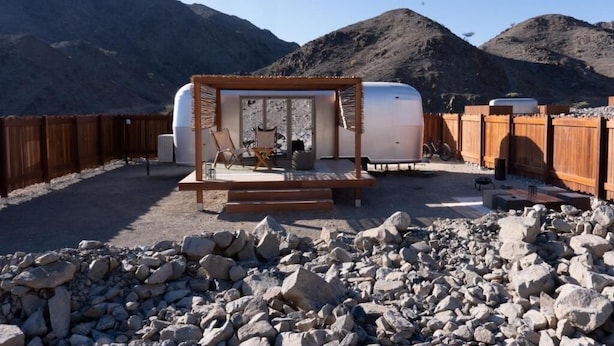 Un véhicule récréatif (VR) doté d’une terrasse en bois et d’une petite scène, entouré d’une clôture en bois et d’un terrain rocheux, avec des montagnes en arrière-plan.