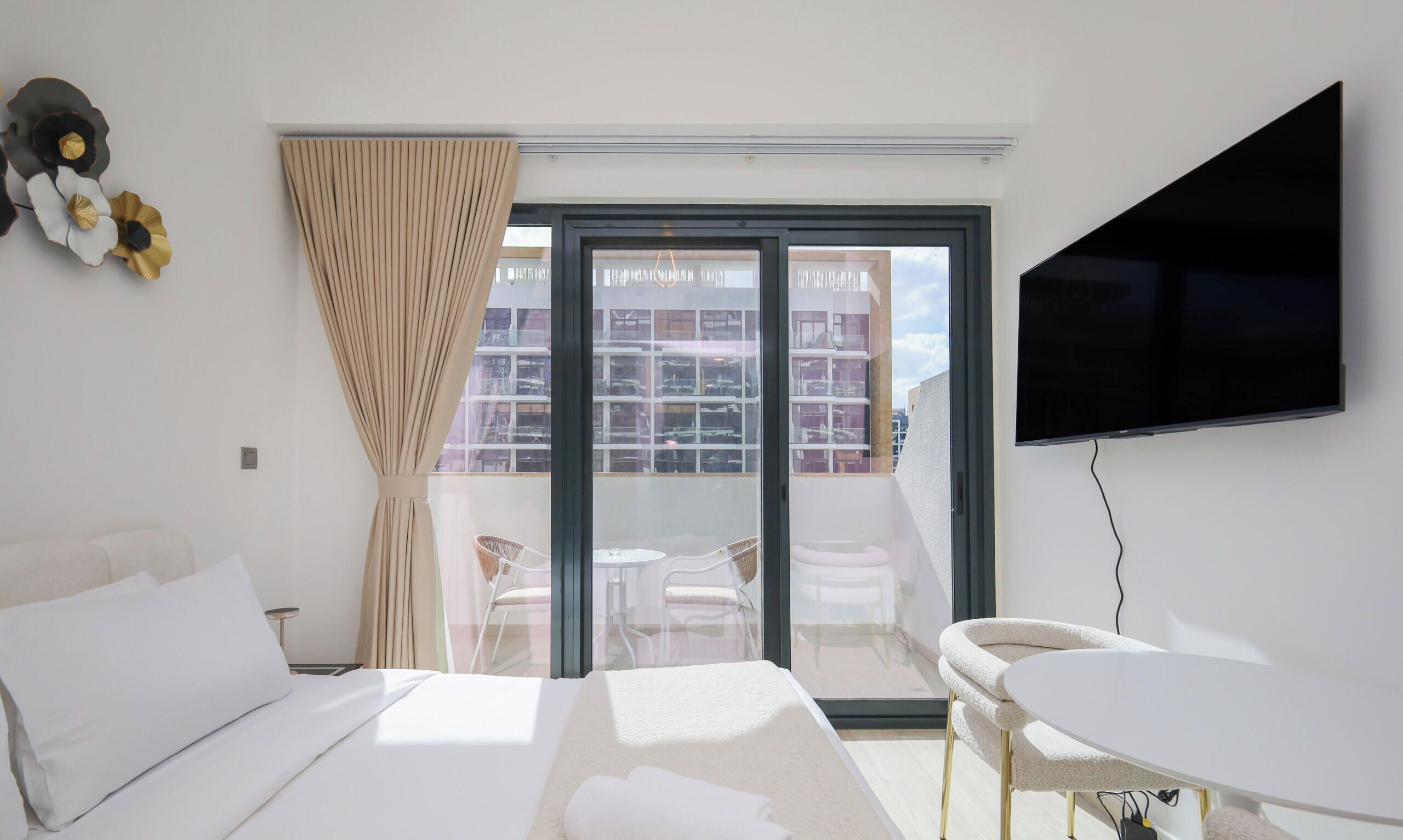 Une chambre d’hôtel comprenant un lit, une télévision, un balcon donnant sur d’autres immeubles et un coin repas avec des chaises et une table.