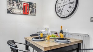Ein Essbereich mit einem Holztisch, gedeckt für zwei Personen, inklusive Tellern, Gläsern, einer Flasche und einer Obstschale. Eine große Wanduhr und ein Stadtbild sind zu sehen.
