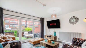 Ein modernes Wohnzimmer mit großem Fernseher, Ledersofas, einem Couchtisch und Blick auf ein rot backsteins Gebäude durch die Glastür.