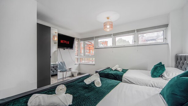 Ein Hotelzimmer mit zwei Betten, einem an der Wand befestigten Fernseher und einem Fenster mit Blick auf Gebäude.