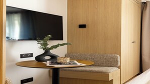 Una habitación moderna con un televisor de pantalla plana montado en la pared, una mesa redonda de madera con un florero y una revista, un banco acolchado y un armario de madera.