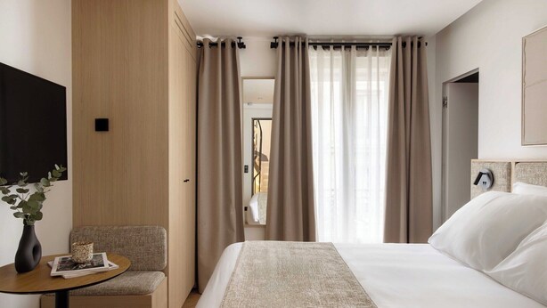 Una habitación de hotel con una cama, una mesita con un florero y una revista, una silla y un televisor montado en la pared.