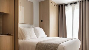 Ein ordentlich bezogenes Bett mit weißen Leinen und einer grauen Tagesdecke, ein Holzschrank und ein großer Spiegel an der Wand.