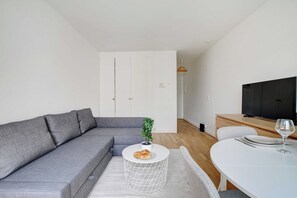 A modern living room with a grey sofa, a round coffee table with a plant, a dining table with plates and a wine glass, and a TV on a wooden stand. - Charming studio - 2P - balcony - Saint Honore (Paris)