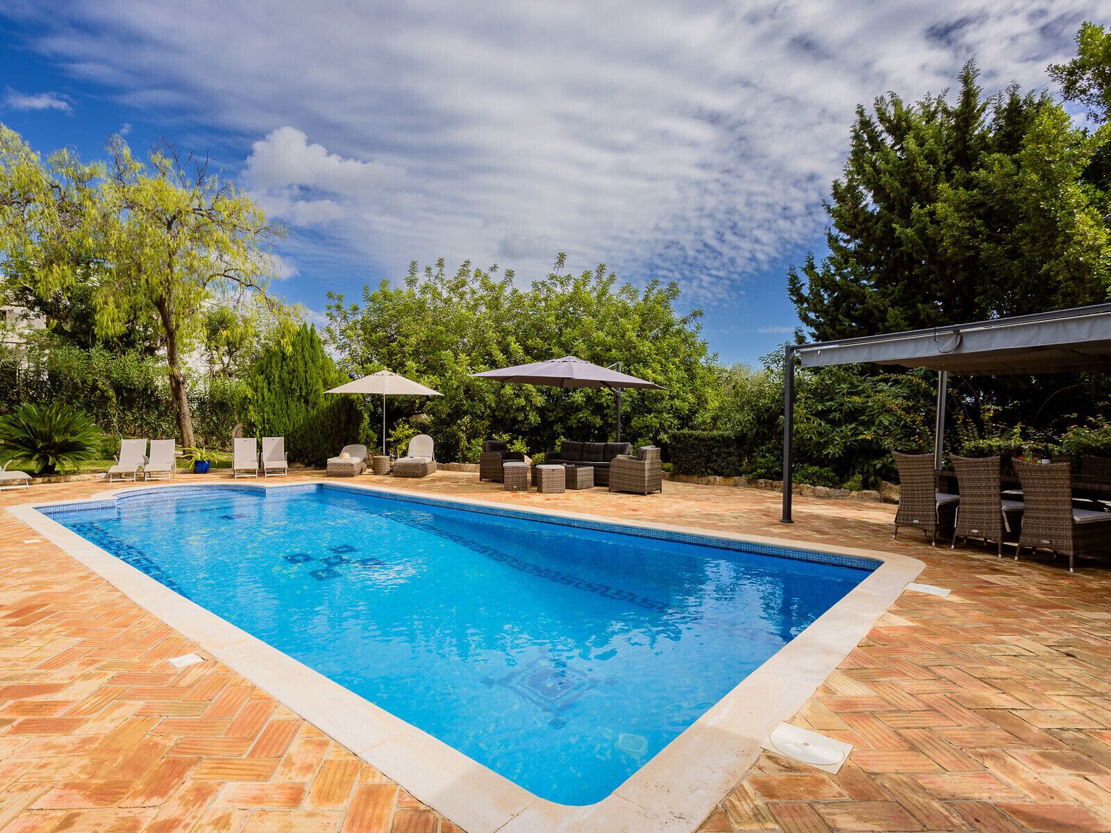 Una piscina rectangular con borde de azulejos, rodeada por un patio con diseño en espiga. Se observan sillones de playa y sombrillas, además de una zona de estar cubierta con mobiliario de mimbre.