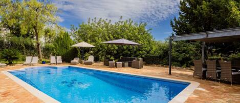Una piscina rectangular con borde de azulejos, rodeada por un patio con diseño en espiga. Se observan sillones de playa y sombrillas, además de una zona de estar cubierta con mobiliario de mimbre.