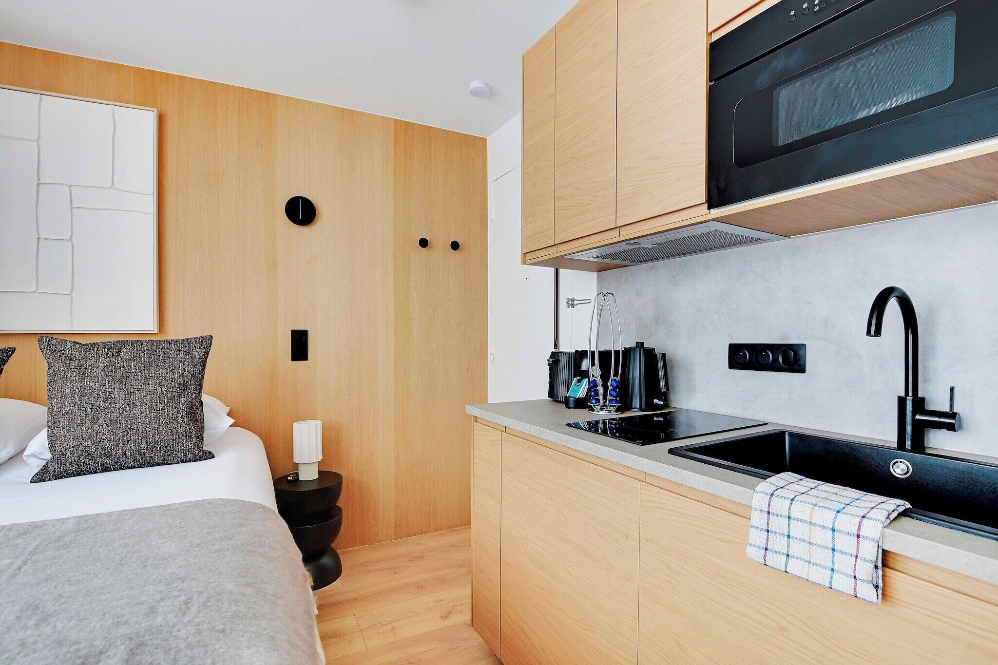 A compact bedroom with a bed, bedside table, and a small kitchenette with a sink and microwave.