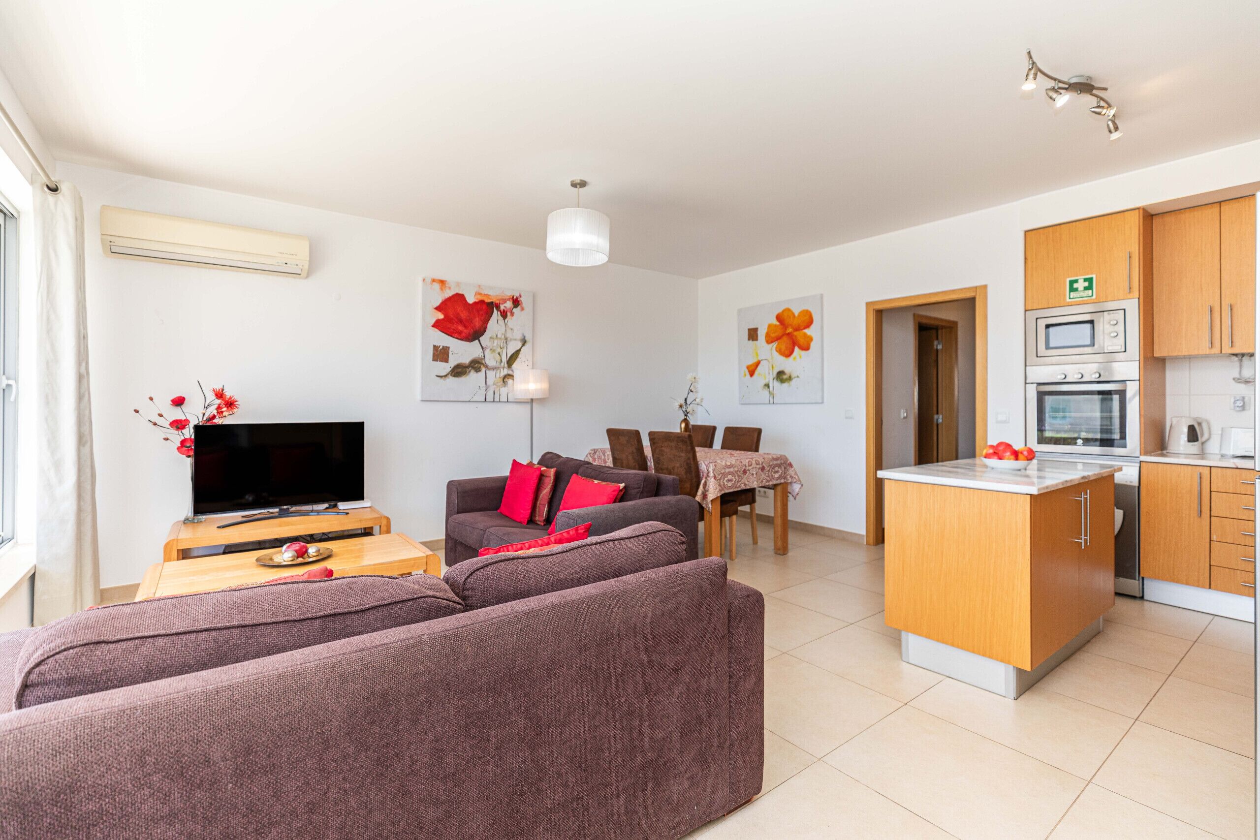 A modern living room with a sofa, TV, and dining area. There is a kitchen with cabinets and appliances.