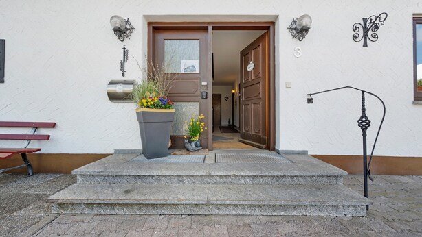 A building entrance with a wooden door, a stone bench, a potted plant, and a wall-mounted light fixture.