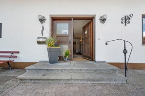Ein Gebäudeeingang mit einer Holztür, einer Steinsitzbank, einem Blumentopf und einer Wandleuchte.