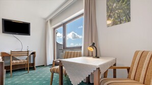 A room with a TV, a table with a lace tablecloth, a sofa, a chair, a window with a view of the sky, and a lamp.